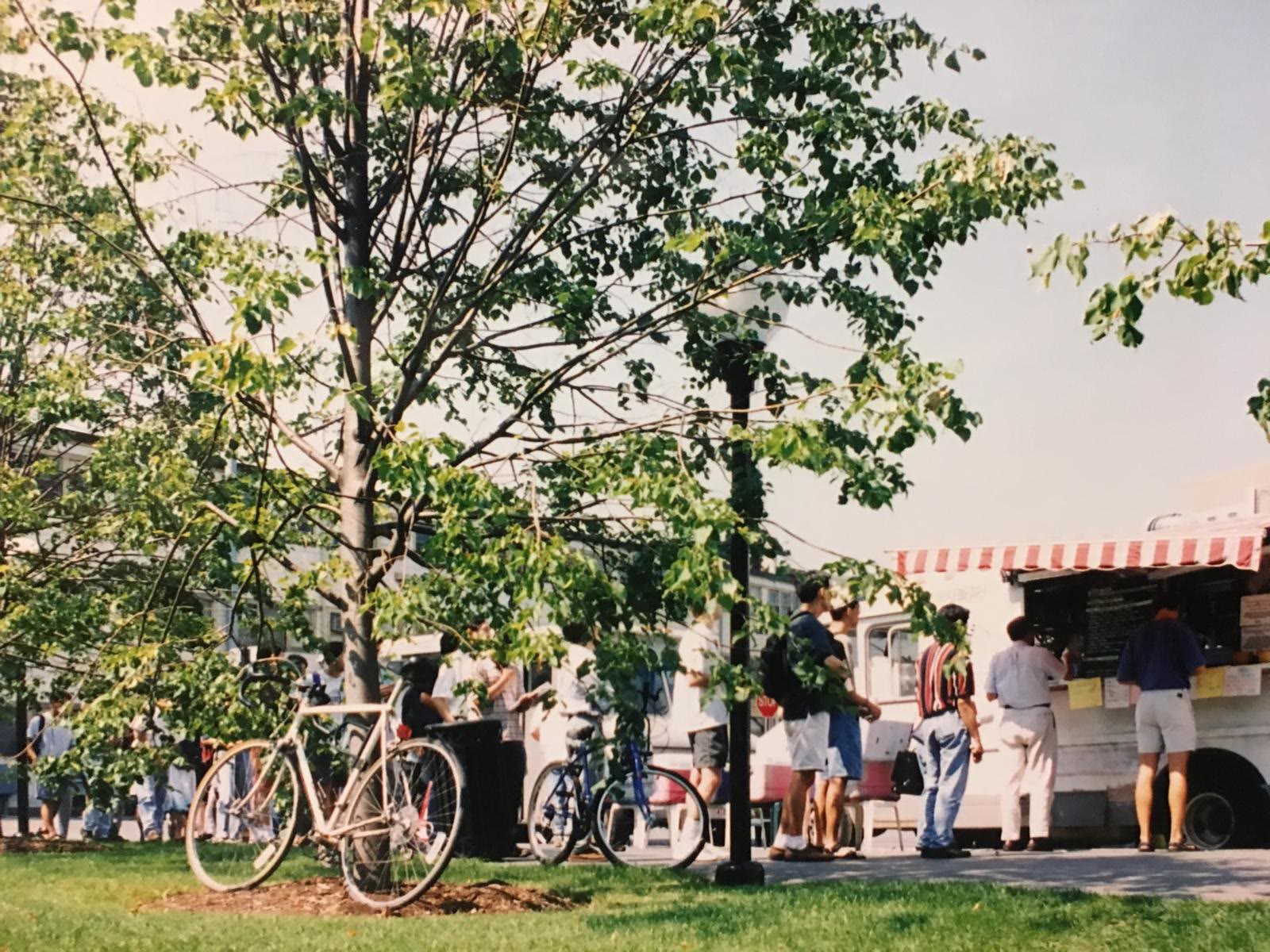 Customers standing at the serving window of the truck, with a red and white striped awning, bicycles leaning against trees nearby.